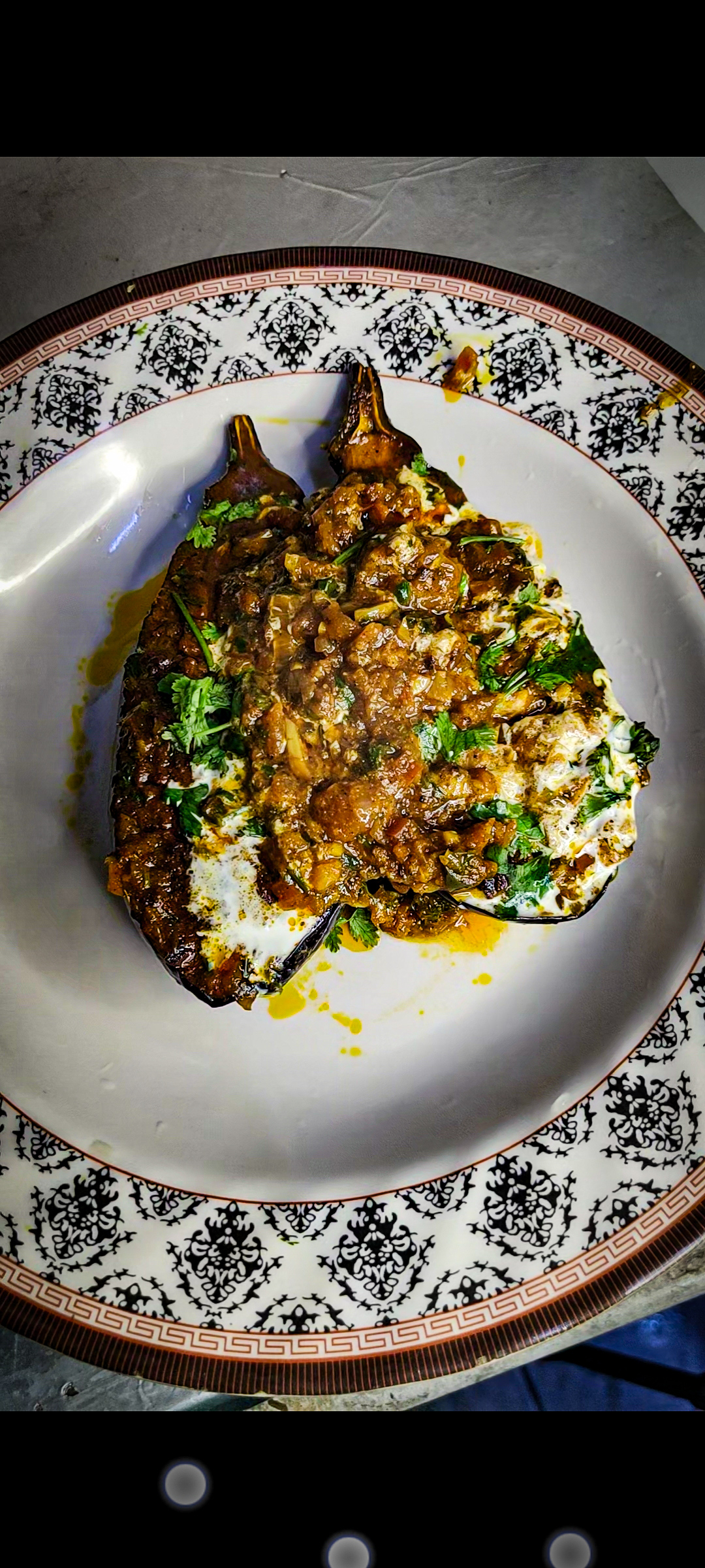 A beautifully plated dish of stuffed and roasted eggplant, topped with a rich, spiced gravy, fresh herbs, and a drizzle of cream, served on an elegant patterned plate — a perfect blend of traditional flavors and artistic presentation.